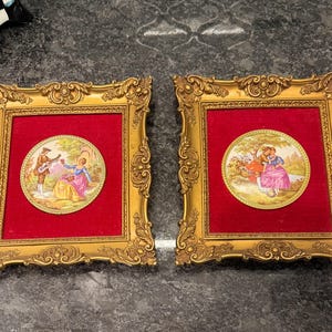 May include: Two ornate gold picture frames with red velvet backing. Each frame holds a circular porcelain plaque depicting a couple in a garden setting. The frames have detailed scrollwork and are displayed on a dark surface.