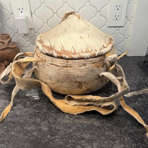May include: A round, lidded container with a textured, fur-like exterior in shades of tan and cream. The lid and body are adorned with long, tan leather strips. The container sits on a dark countertop.