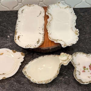 May include: A collection of five antique white porcelain serving dishes with gold trim. The dishes vary in shape, including oval, rectangular, and floral designs. Some feature delicate floral accents, perfect for serving appetizers or desserts.