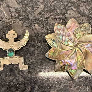 May include: Two decorative brooches. One is a silver-toned figure with a turquoise stone and wing-like details. The other is a flower-shaped brooch with iridescent abalone shell petals and a silver-toned frame. Both are on a dark, textured surface.