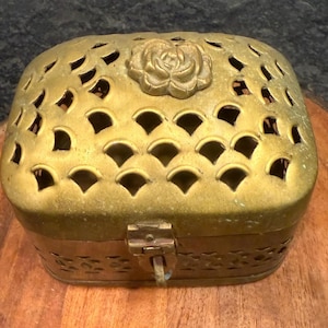 May include: A vintage brass jewelry box with a decorative rose on the lid. The box features intricate cut-out patterns and a clasp closure. The box is approximately 5 inches long, 3 inches wide, and 3 inches tall.