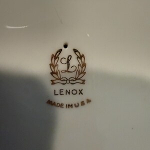May include: Close-up of a white ceramic surface with the Lenox logo in gold. The logo features a stylized "L" within a wreath, with the word "LENOX" below and "MADE IN USA" beneath that.
