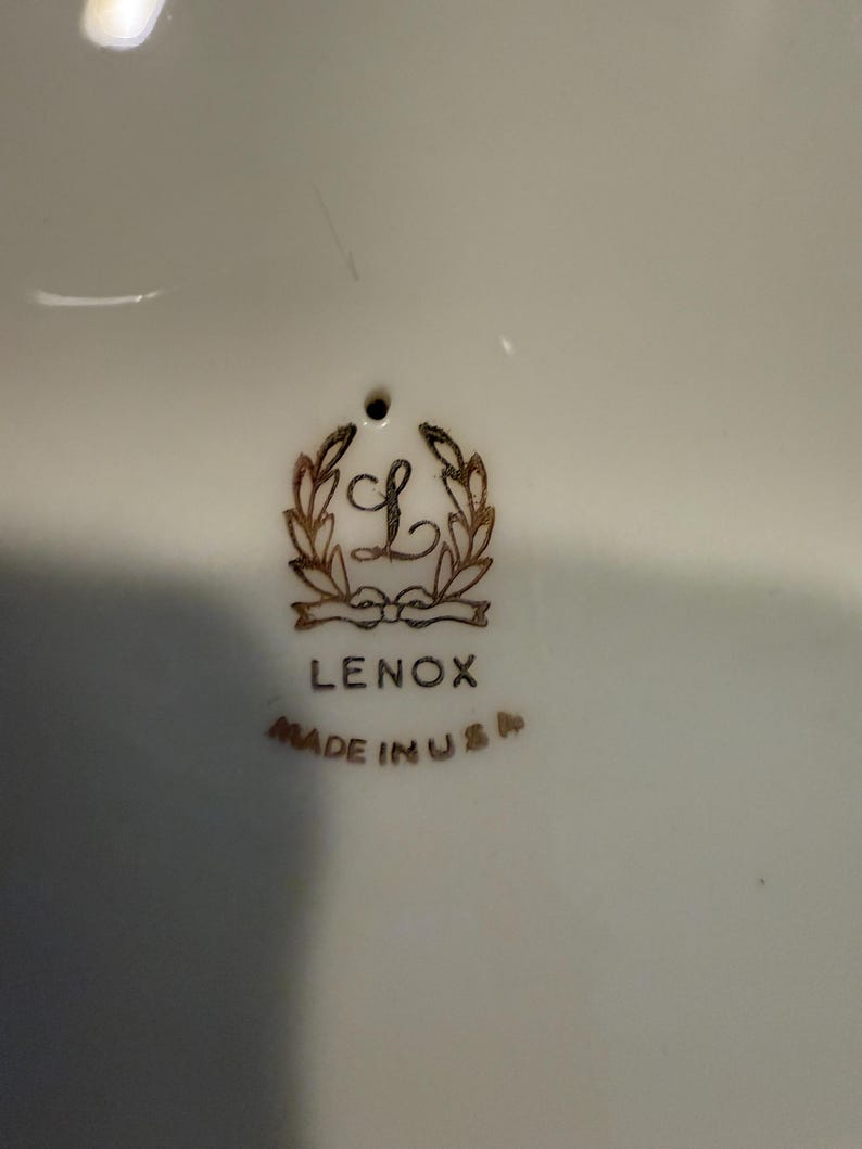 May include: Close-up of a white ceramic surface with the Lenox logo in gold. The logo features a stylized "L" within a wreath, with the word "LENOX" below and "MADE IN USA" beneath that.