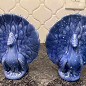 May include: Two blue ceramic peacock figurines. Each peacock has a fanned tail and stands upright. The figurines are a solid blue color and have detailed feather patterns. They are displayed on a dark countertop.