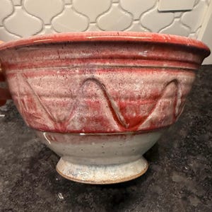 May include: A ceramic mixing bowl featuring a wide rim and a pedestal base. The exterior displays a two-tone glaze with a vibrant red upper section decorated with a raised, wavy line pattern and a smooth, off-white lower section with dark speckles.