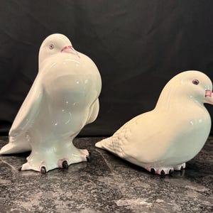 May include: Two white ceramic bird figurines with glossy finishes. One bird stands upright with a puffed chest, while the other rests in a seated position. Both feature small dark eyes, light pink beaks, and dark-tipped feet on their bases.