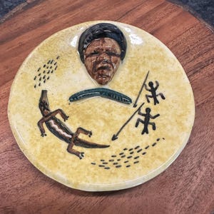 May include: A yellow ceramic plate with a brown and black lizard, two stick figures with spears, and a brown and black face in relief.