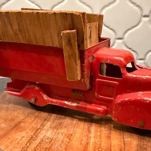 May include: A vintage red toy dump truck with a wooden bed. The truck has a classic design with a detailed front grill and a weathered appearance. It sits on a wooden slice, showcasing its nostalgic charm.
