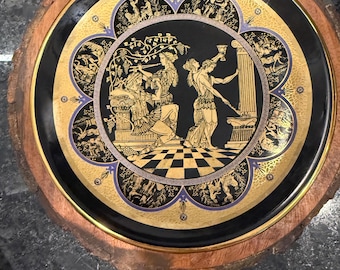 May include: A decorative plate with a black and gold design featuring a classical scene. The plate has a gold rim and is set on a wooden base. The central image depicts figures in classical attire, with a checkered floor.