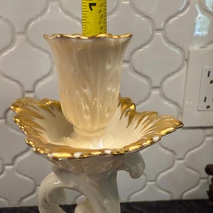 May include: Cream-colored porcelain candle holder with gold trim. The candle holder is approximately 10 inches tall. The base features a decorative, swirling design. The top has a scalloped edge with gold accents.
