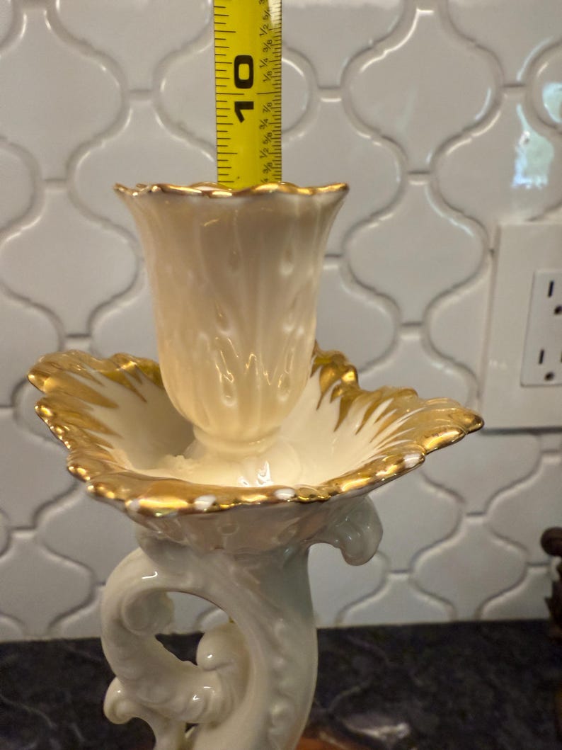 May include: Cream-colored porcelain candle holder with gold trim. The candle holder is approximately 10 inches tall. The base features a decorative, swirling design. The top has a scalloped edge with gold accents.