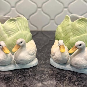 May include: Two ceramic duck figurines with yellow beaks and white feathers, sitting on a light blue base. Behind them are two cabbage-shaped containers in shades of green. The figurines are on a dark surface.