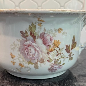 May include: White ceramic chamber pot with a floral design featuring pink and purple roses, yellow accents, and green leaves. The pot has a handle and a decorative rim with a gold-colored trim.