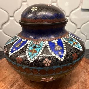 May include: A decorative, lidded jar with intricate enamel work. The jar features a dark blue base, a black body with floral and geometric patterns in blue, white, and red, and a dark blue lid with white floral accents.