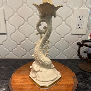 May include: A cream-colored ceramic candlestick holder with gold trim. The base is a stylized seashell shape, and the stem features a dolphin design. The top has a fluted cup for holding a candle. The candlestick is displayed on a wooden round.