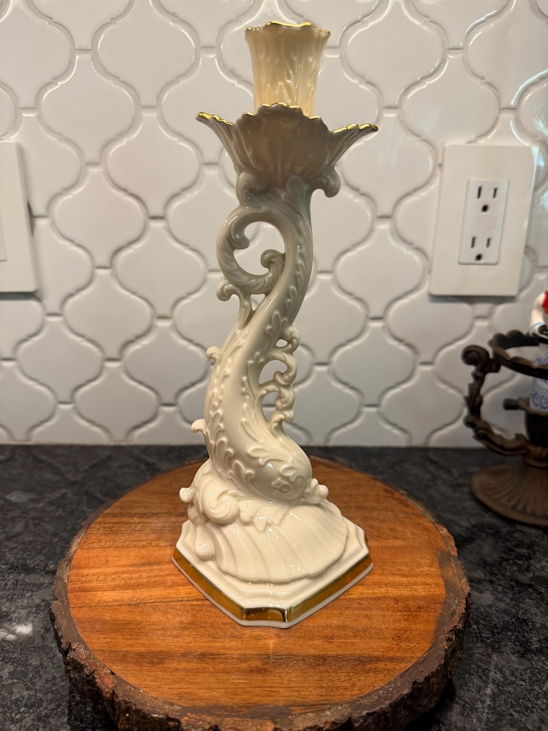May include: A cream-colored ceramic candlestick holder with gold trim. The base is a stylized seashell shape, and the stem features a dolphin design. The top has a fluted cup for holding a candle. The candlestick is displayed on a wooden round.