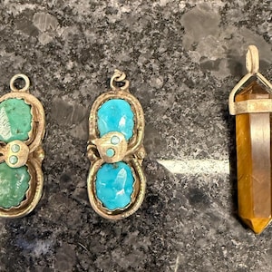 May include: Three pendants with gemstone accents. Two pendants feature turquoise stones in a gold-colored setting. The third pendant has a rectangular, amber-colored stone with silver-colored metal accents. The pendants are displayed on a dark, speckled surface.