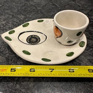 May include: A white ceramic egg cup with green and orange polka dots, sitting on a white ceramic saucer with green polka dots. The saucer has a black circle with the text "Devon Barn Pottery" in gold lettering.