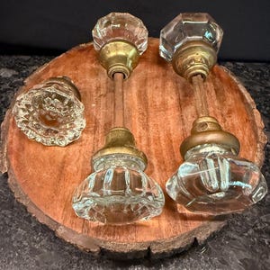 May include: A set of three clear glass door knobs with faceted surfaces and brass-toned metal bases. Two of the knobs are attached to long metal spindles, while one knob is detached, showing the intricate glass pattern and metal hardware.