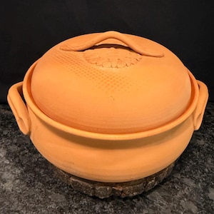 May include: A round, terracotta-colored clay cooking pot with a matching lid. The lid features a central floral-shaped relief and a curved handle. The pot has two side handles and a smooth, matte finish, showcasing a natural, earthy orange tone.