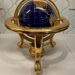 May include: A decorative globe featuring a deep blue ocean surface with inlaid stone continents in various colors. The globe is mounted within a polished gold-tone metal meridian ring and supported by a three-legged stand with ornate feet.