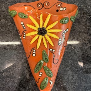 May include: Orange ceramic wall pocket with a sunflower design. The flower has yellow petals and a black center. Green leaves and white dots are painted on the orange surface. Black swirls and accents complete the design.