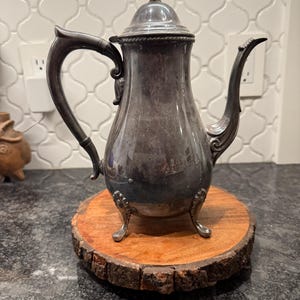 May include: A silver-colored antique coffee pot with a curved handle and spout, resting on a round wooden base. The pot has a decorative lid and stands on three small feet. The wooden base has a natural, rustic edge.