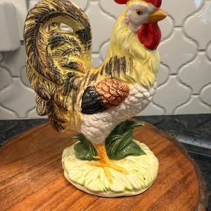 May include: A ceramic rooster figurine with a yellow body, red comb, and black and brown accents. The rooster stands on a green and yellow base with green leaves. The figurine is displayed on a wooden tray.