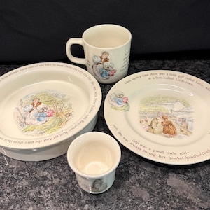May include: A ceramic dinnerware set featuring Peter Rabbit illustrations and text. The set includes a mug, a small cup, a bowl, and a plate. Each piece is off-white with colorful storybook scenes and printed text from the Beatrix Potter tales.