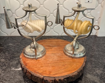 May include: Two antique silver candle holders with spiral candles. Each holder has a decorative frame, a small chain, and a conical pendant. The candle holders are on a round wooden base with a natural edge.