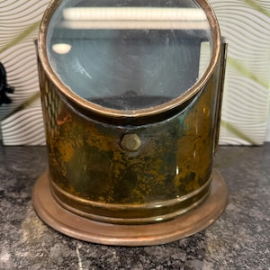 May include: A vintage brass magnifying glass with a circular lens and a cylindrical base. The brass has a weathered patina, and the lens is set in a brass frame. The magnifying glass sits on a round base. A small brass handle is at the top.
