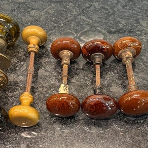 May include: A collection of antique door knobs in various styles and colors. The knobs feature glass, wood, and metal components, with shades of brown, amber, and gold. Some have ornate detailing, while others are simpler in design.