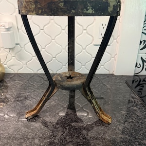 May include: A small, three-legged metal stand featuring a circular top with a distressed, weathered green and brown finish. The structure includes black metal legs that converge at a central base plate, which is accented with a cutout star shape.
