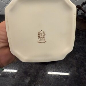 May include: Off-white ceramic vase base with a unique octagonal shape. The bottom features the Lenox logo and the text "MADE IN U.S.A." in brown lettering. The vase is set against a dark, textured surface.
