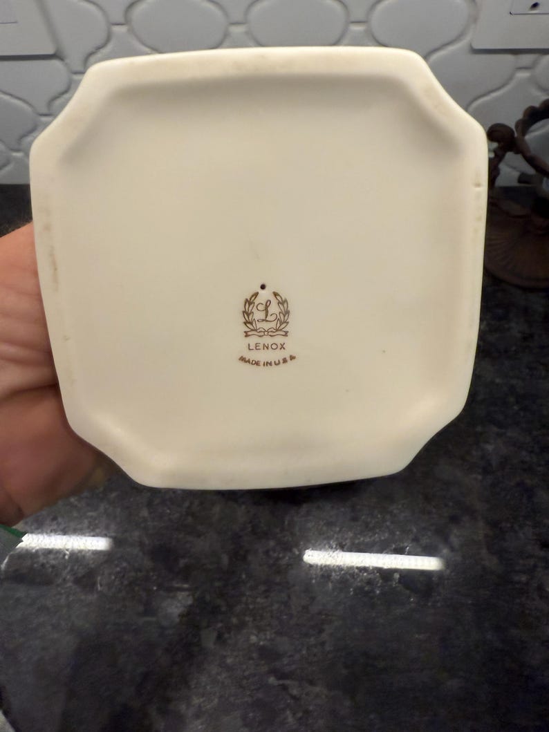 May include: Off-white ceramic vase base with a unique octagonal shape. The bottom features the Lenox logo and the text "MADE IN U.S.A." in brown lettering. The vase is set against a dark, textured surface.