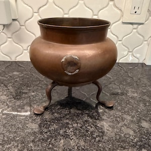 May include: A round copper pot with a wide opening and a smooth, metallic finish. The vessel features three curved legs extending from the base and a small, circular, silver-toned patch or rivet detail on the front side of the exterior body.
