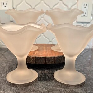 May include: Four frosted glass dessert dishes with a ruffled rim and pedestal base. The dishes are arranged on a wooden serving board. The glass has a soft, diffused appearance, and the overall aesthetic is delicate and vintage.