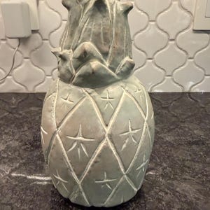 May include: A decorative pineapple figurine with a weathered, pale green finish. The surface features a diamond-patterned texture with carved star-like details within each section and a pointed, leafy top section with a muted, earthy patina.