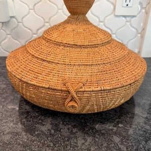 May include: A woven basket with a lid, featuring a conical handle. The basket is made of light brown wicker and has a textured surface. The lid is secured with a woven closure. The basket is likely used for storage or decoration.