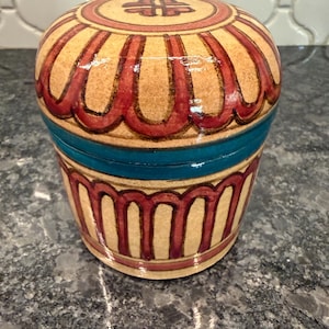 May include: A decorative, hand-painted ceramic jar with a lid. The jar features a tan base with red and teal accents. The lid has a red border and a central design. The jar is approximately 5 inches tall and 5 inches wide.