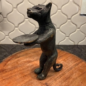 May include: A bronze cat figurine holding a small, round tray. The cat is standing upright with its tail curled. The figurine has a weathered, antique appearance and is set on a wooden surface.