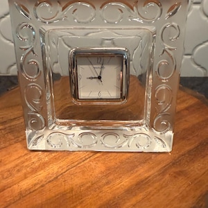 May include: A clear glass square clock with a silver-toned clock face. The clock face has black hands and Roman numerals. The clock is set in a decorative glass frame with an etched swirl design. The clock is sitting on a wooden surface.