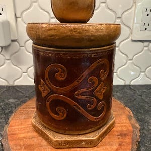 May include: A cylindrical brown container with a textured surface featuring embossed scrollwork patterns. The lid is topped with a spherical knob, and the base is hexagonal with a light tan border. The finish appears aged with dark brown tones.