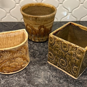 May include: Three handcrafted ceramic planters in earthy tones. The set includes a cylindrical pot, a square planter with a floral pattern, and a half-cylinder planter with a carved design. These decorative pots are suitable for indoor or outdoor use.