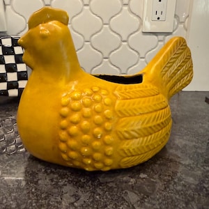 May include: A ceramic hen-shaped planter in a vibrant yellow hue. The planter features a textured body with a detailed tail and a crest. The opening is at the top, and the planter is set on a dark countertop.