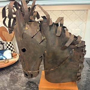 May include: A rustic metal sculpture of a horse's head, featuring a textured, weathered appearance. The sculpture is mounted on a small, rectangular wooden base. The horse's mane and facial features are crafted with intricate detail, showcasing a unique artistic style.