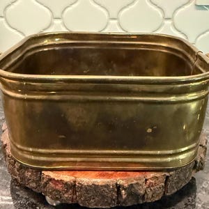 May include: A rectangular brass basin with rounded corners and a polished metallic finish. The container features a smooth, slightly reflective surface with subtle signs of wear and small handles on the sides, showcasing a classic metal construction.