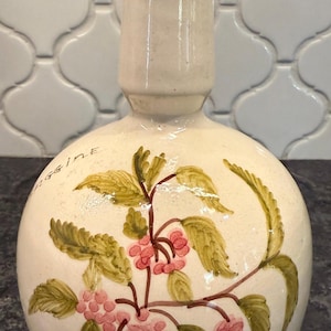 May include: Cream-colored ceramic bottle with a round body and a cylindrical neck, adorned with a hand-painted floral design featuring pink berries and green leaves. The neck has two brown stripes. The bottle is likely a decorative piece.