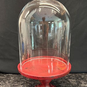 May include: A clear glass cloche dome with a spherical handle sits atop a round, distressed red pedestal stand. The stand features a decorative, ornate base with black accents, providing a sturdy support for the transparent glass cover.