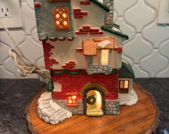 May include: A ceramic Christmas village house with a red brick facade, green trim, and a stone chimney. The house has lit windows and a small wreath over the arched doorway. It sits on a round wooden base.